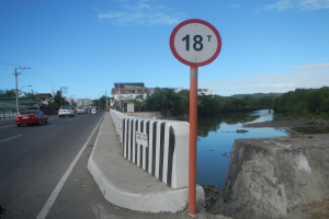 Ein Geschwindigkeitsbegrenzungsschild am Straßenrand neben einem Fluss, mit Fahrzeugen, einer Barriere, Bäumen, Gebäuden, Strommasten mit Drähten und einem bewölkten Himmel im Hintergrund.