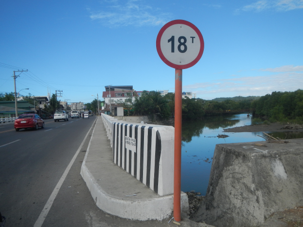 Ein Geschwindigkeitsbegrenzungsschild am Straßenrand neben einem Fluss, mit Fahrzeugen, einer Barriere, Bäumen, Gebäuden, Strommasten mit Drähten und einem bewölkten Himmel im Hintergrund.