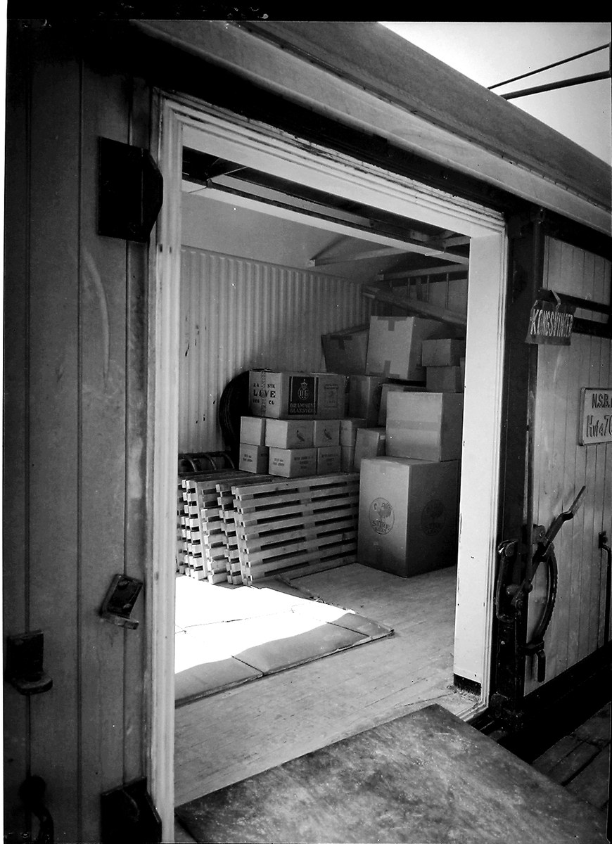 Schwarze und weiße Fotografie eines Metall-Shipping-Containers mit einer Tür auf der linken Seite, gefüllt mit ordentlich gestapelten Kartons.