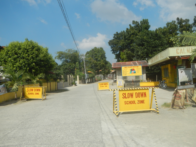 Eine Straße mit einem gelben "Slow Down School Zone"-Schild auf der linken Seite, parkende Fahrzeuge auf der rechten Seite und einem Hintergrund mit Bäumen, Gebäuden, Polen, Drähten und dem Himmel.