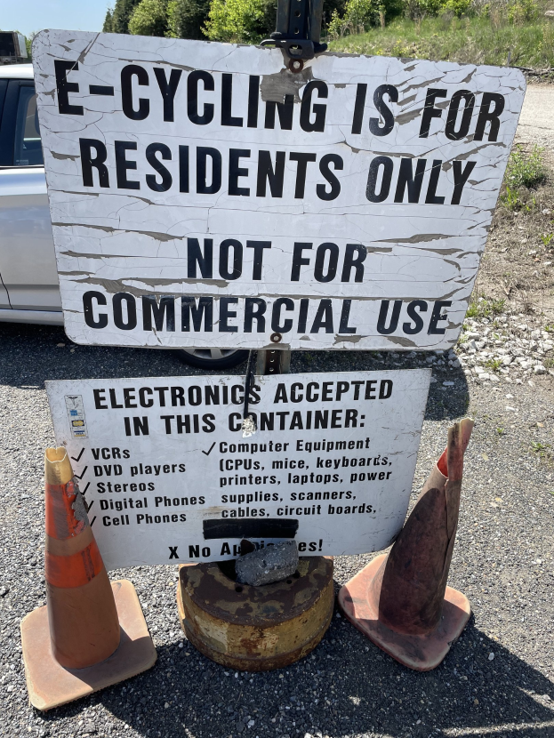 Schild mit Aufschrift "e-Bike-Fahren nur für Anwohner" neben einer Straße mit Absperrbaken, einem Fahrzeug, Bäumen und einem klaren blauen Himmel im Hintergrund.