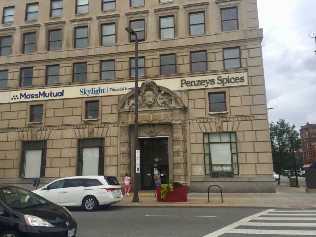 Großes Gebäude mit parkenden Autos davor, Straßenlaternen, Verkehr, Fußgänger auf dem Gehweg, Grünfläche und bewölkter Himmel, identifiziert als Hauptsitz der Massachusetts Mutual Financial Group.