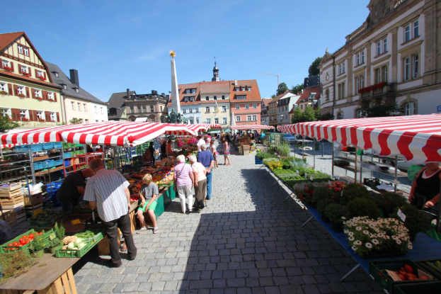 Ein belebter Markt im alten Stadtkern von Heidelberg mit Menschen, die spazieren gehen, auf Bänken sitzen und in der Nähe von Zelten stehen, mit Gemüsekörben auf Tischen, umgeben von Gebäuden, Bäumen und einem klaren blauen Himmel.