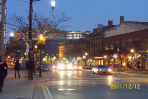 Eine nächtliche Stadtstraße voller Verkehr, beleuchtet von Straßenlaternen, mit Autos, Fußgängern, Gebäuden, Bäumen und einem sternenklaren Himmel im Hintergrund, mit einem Wasserzeichen auf der rechten Seite.