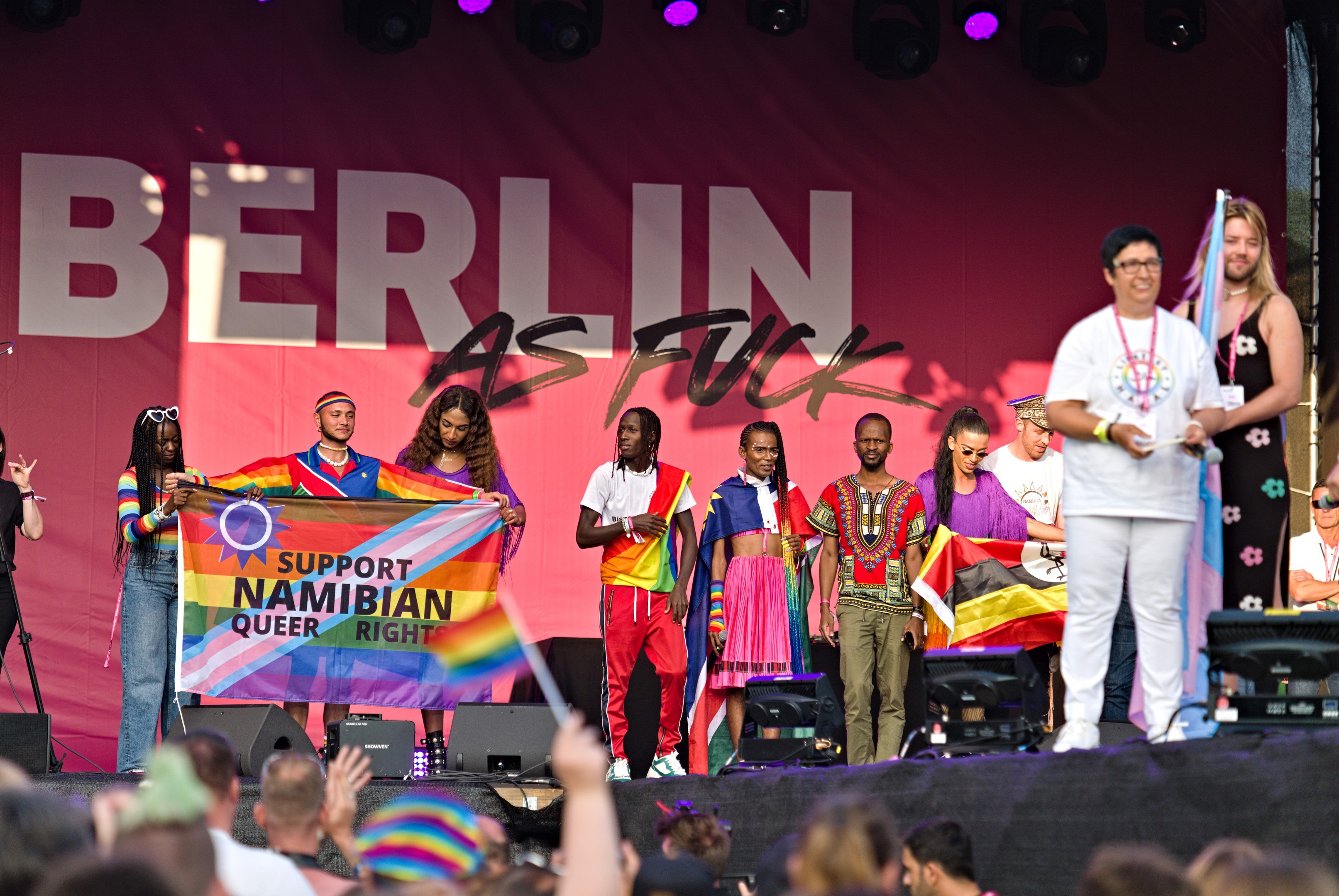 Eine Gruppe von Menschen auf einer Bühne mit einem Banner, auf dem 'Berlin as Fuck' steht, mit Lautsprechern, Mikrofonen und einer Menge mit erhobenen Händen darunter.