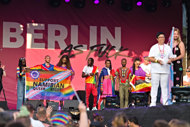 Eine Gruppe von Menschen auf einer Bühne mit einem Banner, auf dem 'Berlin as Fuck' steht, mit Lautsprechern, Mikrofonen und einer Menge mit erhobenen Händen darunter.