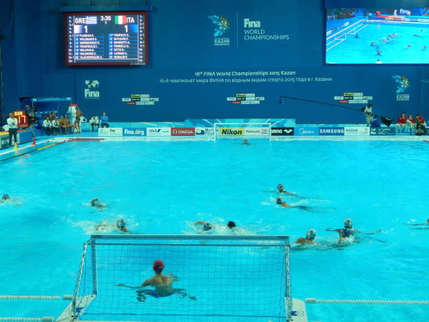 Eine Gruppe von Menschen, die Wasserball in einem großen Pool mit einem Netz in der Mitte spielen, Zuschauern auf der linken Seite und einem Bildschirm mit Text und Logos im Hintergrund bei den FINA-Weltmeisterschaften 2016 in Kazan.