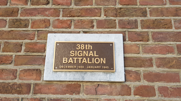 Eine Ziegelwand mit einem rechteckigen Schild, auf dem in fetter, weißer Schrift "38. Signalbataillon" steht.