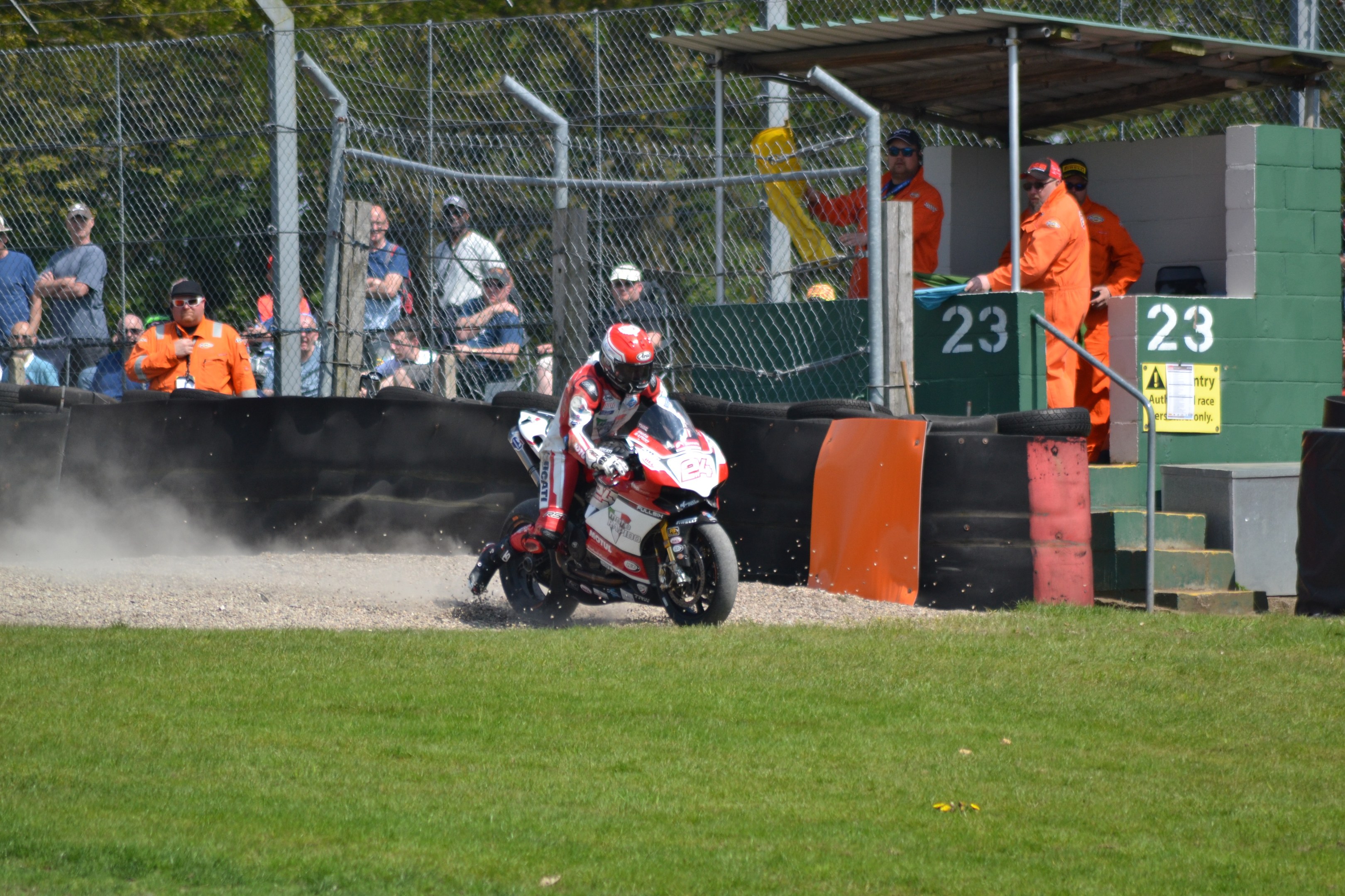 Eine Person mit Helm und Handschuhen fährt ein Motorrad über ein grasbewachsenes Feld, mit Zuschauern, Absperrungen, Treppen und einem Schuppen im Hintergrund.