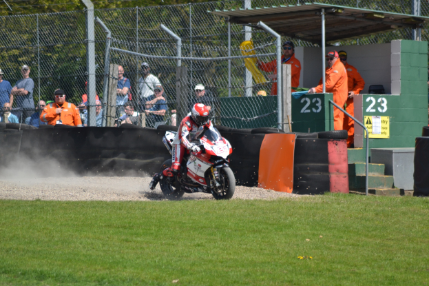 Eine Person mit Helm und Handschuhen fährt ein Motorrad über ein grasbewachsenes Feld, mit Zuschauern, Absperrungen, Treppen und einem Schuppen im Hintergrund.