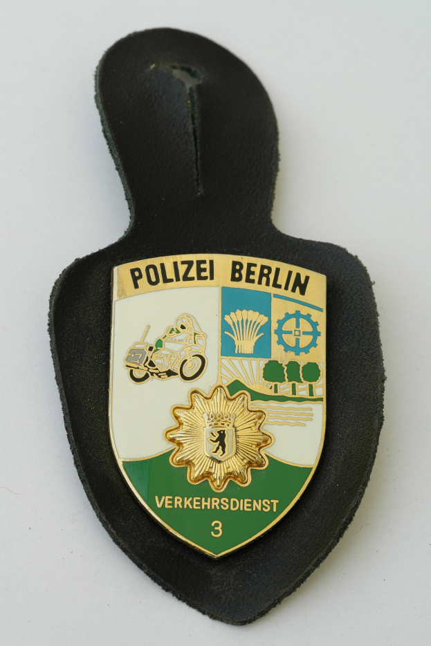 Ein schildförmiges Polizeibadgebnis mit "Polizei Berlin" in schwarzer fetter Schrift auf einem blauen Hintergrund mit einem weißen Stern und Rand, das auf einer Oberfläche ruht.