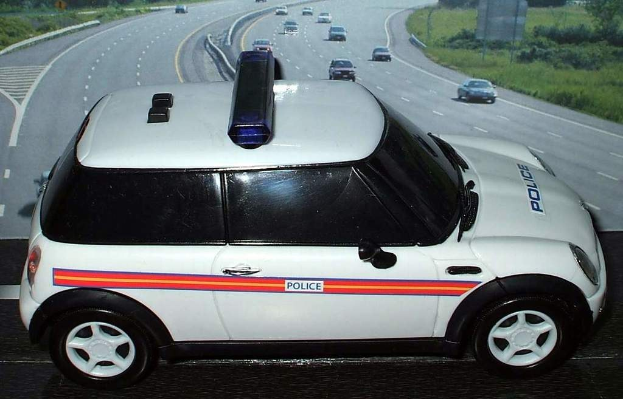 Weißer Polizeiwagen am Straßenrand mit vorbeifahrendem Verkehr, Gras und Bäumen auf beiden Seiten, aufgenommen außerhalb einer Stadt.