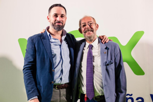 Zwei M├Ąnner in Anz├╝gen l├Ącheln nebeneinander vor einer Wand, wobei der Mann rechts eine Brille tr├Ągt; ein Schild im Hintergrund zeigt "Vodafone und Vodacom ├╝bernehmen Partnerschaft."