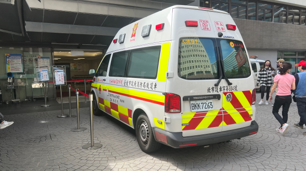 Ein Rettungswagen steht vor einem Gebäude mit Menschen auf der rechten Seite, eine Person sitzt auf einem Stuhl im Hintergrund und Schilder mit Text und Plakaten an der linken Wand, auf denen "Ambulance Service in Hong Kong" steht.