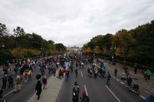 Eine große Gruppe von Menschen marschiert eine baumbestandene Straße mit Laternenpfählen entlang, mit Kameras in der Hand, mit einem Gebäude und einem klaren Himmel im Hintergrund, was auf eine Demonstration in Berlin hinweist.