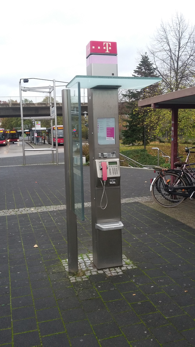 Ein Münzfernsprecher steht an der Straße neben einer Bushaltestelle, daneben parkt ein Fahrrad; im Hintergrund sind Bäume, Fahrzeuge auf der Straße, eine Brücke und ein klarer blauer Himmel zu sehen.