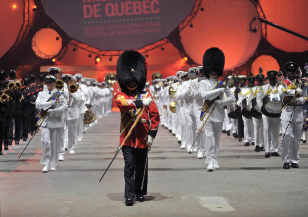 Eine Gruppe uniformierter Militärpersonen marschiert im Gleichschritt, einige spielen Instrumente, während der Eröffnungszeremonie des Montreal International Festival of Military Bands.