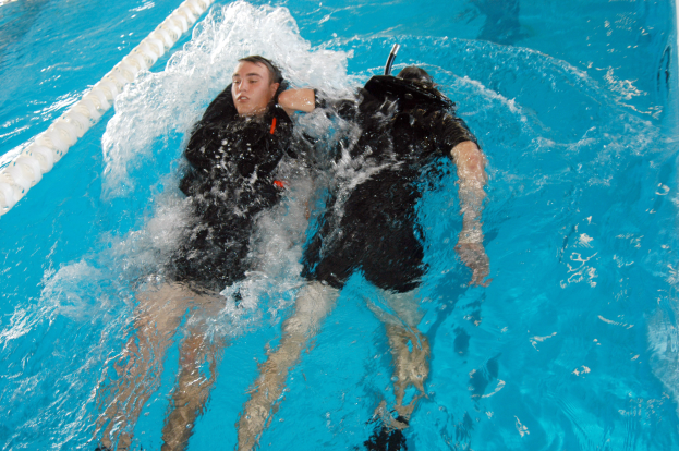 Zwei Personen in schwarzen Neoprenanzügen und Tauchausrüstung, eine mit einem Sauerstoffzylinder, führen einen Rückenschwimm in einem Schwimmbad durch.