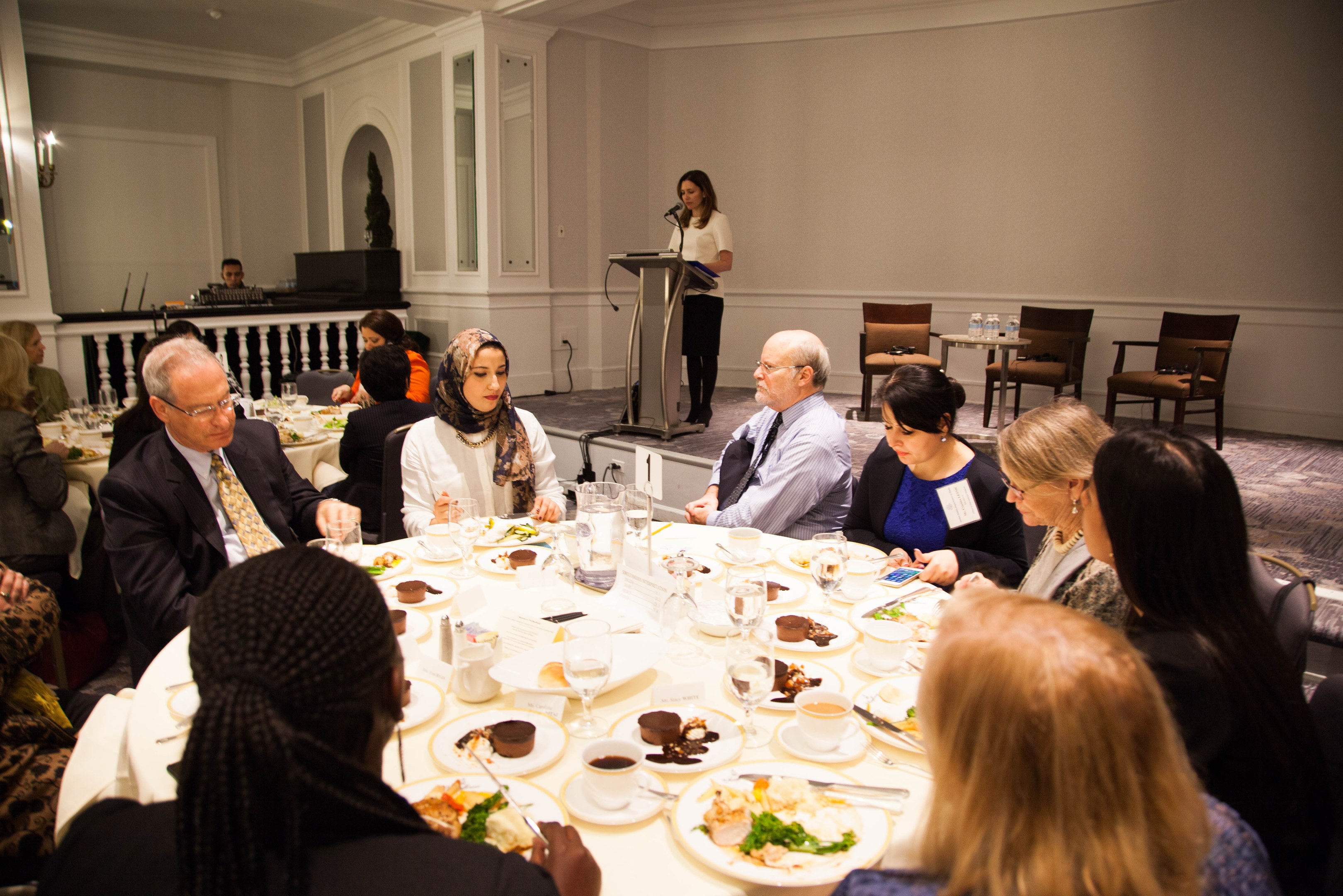 Gruppe von Menschen, die um einen Esstisch mit Essen und Besteck sitzen, in einem Raum mit einer Frau an einem Podium und einer anderen Person in der Nähe eines Geländers.
