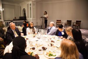 Gruppe von Menschen, die um einen Esstisch mit Essen und Besteck sitzen, in einem Raum mit einer Frau an einem Podium und einer anderen Person in der Nähe eines Geländers.