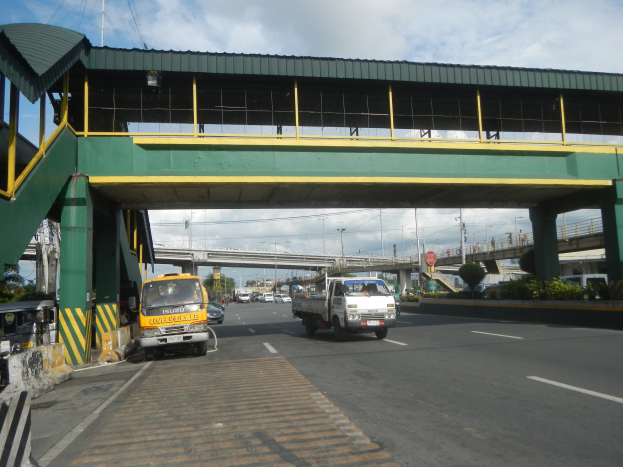 Mautstelle mit Fahrzeugen auf einer Straße unten, eine Brücke mit Geländern und Pfeilern, Laternenmasten, Schilder, Bäume und ein bewölkter Himmel im Hintergrund, mit einem Lastwagen, der neben der Brücke fährt.