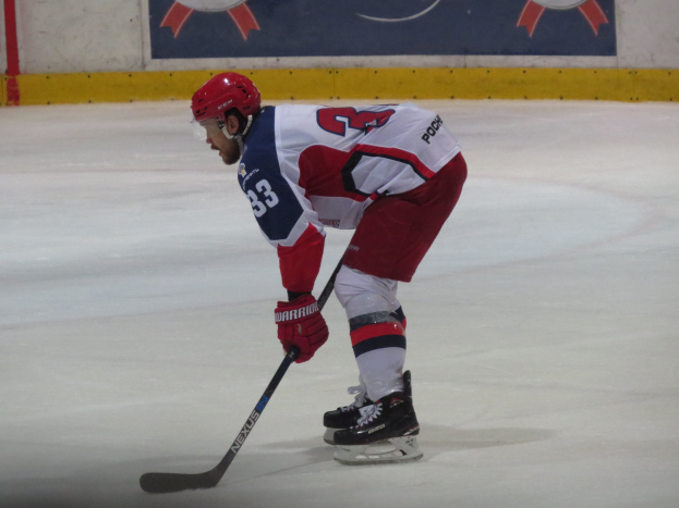 Eishockey-Spieler in voller Ausrüstung auf dem Eis mit einem Stock, der einer Bannerwand gegenübersteht.