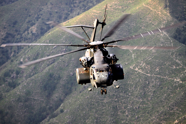 Milit├Ąrhelikopter über ein Gebirgsmassiv mit saftigen grünen Hügeln im Hintergrund mit ausgefahrenem Fahrwerk.