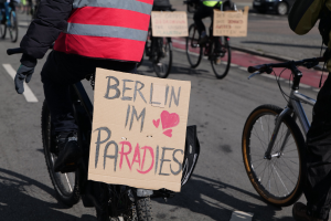 Eine Gruppe von Menschen, die auf Fahrrädern eine Straße entlangfährt, mit einem "Berlin I'm Paradies"-Schild im Vordergrund und einem Auto im Hintergrund, das Bild leicht unscharf.