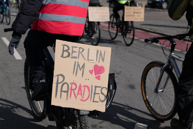 Eine Gruppe von Menschen, die auf Fahrrädern eine Straße entlangfährt, mit einem "Berlin I'm Paradies"-Schild im Vordergrund und einem Auto im Hintergrund, das Bild leicht unscharf.