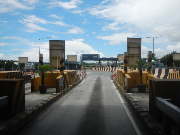 Straße mit einer Mautstelle, Absperrpfählen, Verkehrskegeln, Topfpflanzen, Hinweisschildern, Straßenmasten, Bäumen, einer Brücke und einem bewölkten Himmel.