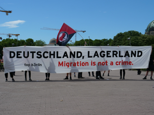 Eine Gruppe von Menschen mit einem Banner 'Deutschland, Lagerland Migration ist kein Verbrechen' und einer Flagge, im Hintergrund Straßenlaternen, Bäume, Kräne, ein Gebäude und ein bewölkter Himmel.