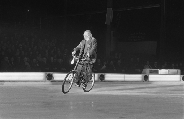 Schwarz-weißes Bild eines Mannes, der auf einem Eisstadion Fahrrad fährt, mit einer Gruppe von Menschen im Hintergrund und einer Wand dahinter.