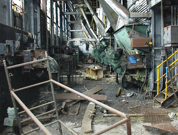 Ein altes, ungenutztes Fabrikinnere mit Maschinen, Schutt auf dem Boden, Fenstern und Geländern auf beiden Seiten.