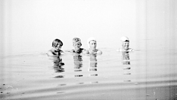 Schwarz-weiß-Foto von vier Kindern, die in einem Pool schwimmen und alle Schwimmkappen tragen.