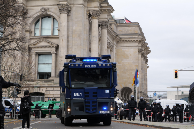 Eine Gruppe von Polizisten steht vor einem großen Gebäude mit Fenstern, Säulen und Bögen, mit Fahrzeugen auf der Straße und einer Person, die eine Kamera hält. Bäume, Ampeln, Fahnenmasten und ein klarer blauer Himmel sind im Hintergrund zu sehen.