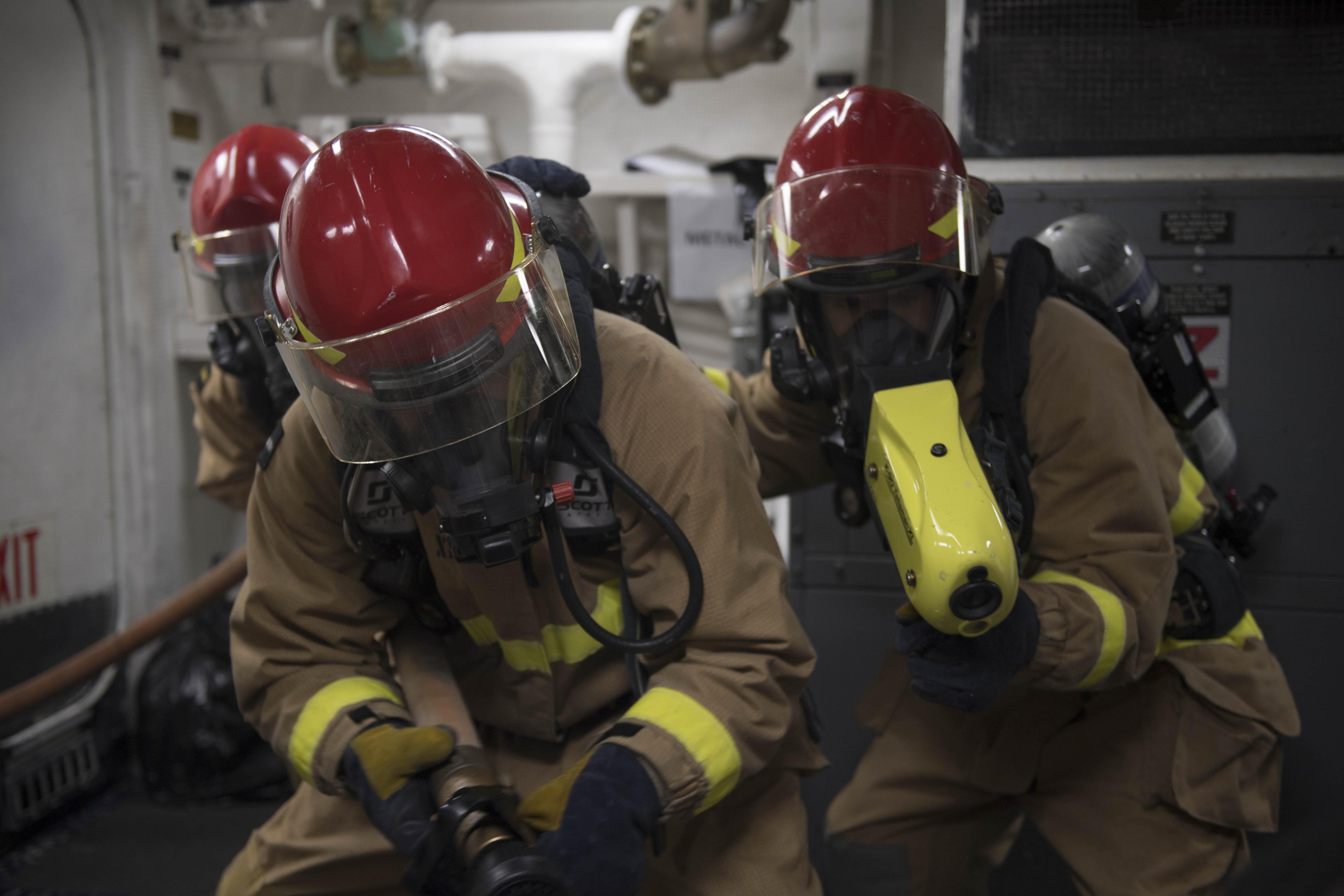 Feuerwehrleute mit Helmen und Gasmasken arbeiten an einem Feuerhydranten in der Nähe eines Ausgangsschilds, mit Rohren und Equipment im Hintergrund.
