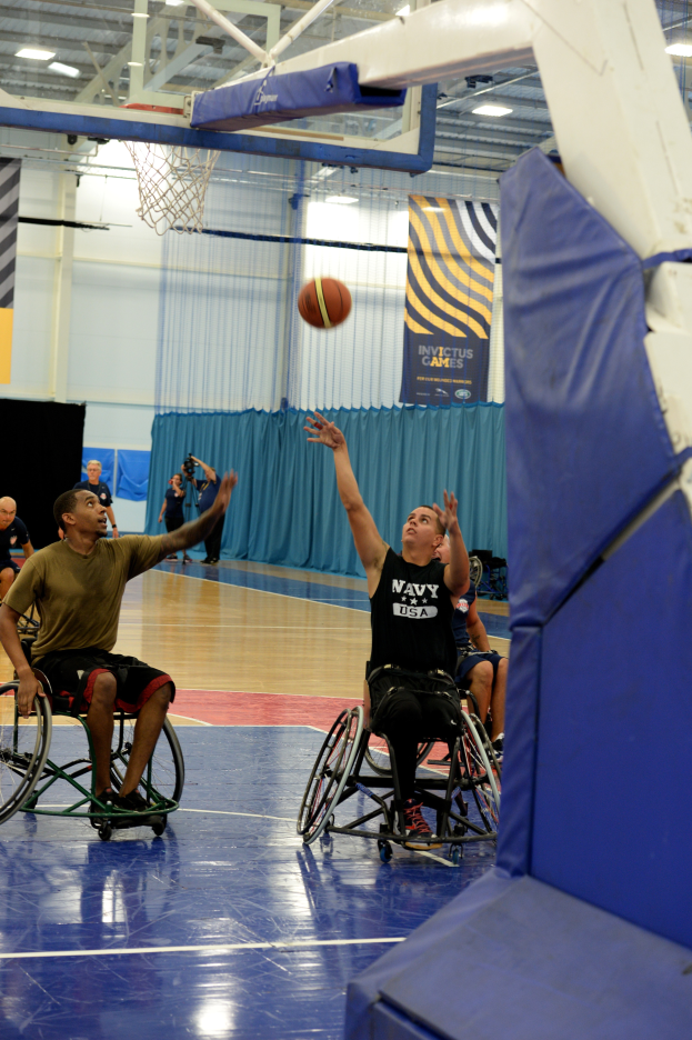 Eine Gruppe von Menschen in Rollstühlen spielt Basketball in einer Turnhalle mit einem Basketballkorb, Bannern mit Text und Deckenbeleuchtung.