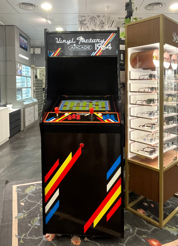 Ein Geschäft mit einem zentralen Vintage-Arcade-Automaten, einer Glasvitrine auf der rechten Seite und Retro-Beleuchtung mit Schränken, Fernseher und anderen Gegenständen im Hintergrund.