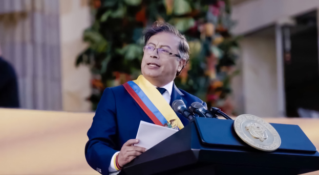 Ein Mann in einem blauen Anzug und Krawatte steht an einem Podium, hält Papiere und spricht in ein Mikrofon, mit einer Pflanze und einer Wand dahinter, was auf eine formelle Umgebung hinweist.