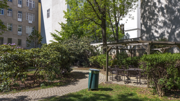 Ein Park mit einem grünen Müllcontainer in der Mitte, umgeben von Gras, Pflanzen, Bäumen, Gebäuden mit Fenstern, verstreuten Stühlen und Tischen sowie einer Hütte im Hintergrund.