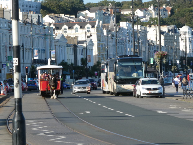 Eine belebte Stadtstraße mit hohen Gebäuden, Fahrzeugen, Fußgängern, Verkehrszeichen, Laternen, Schildern, Bäumen und einem klaren blauen Himmel.