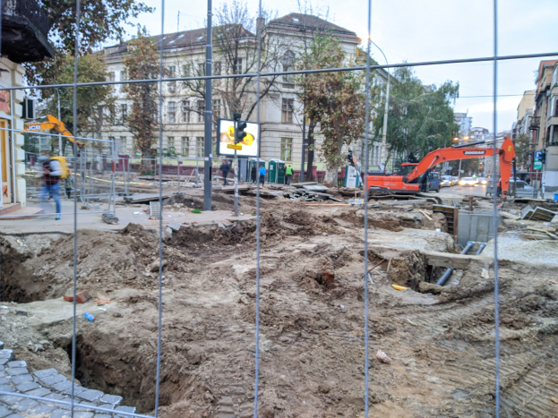 Baustelle mit großem Erdaushub umgeben von einem Metallzaun, angrenzende Straße mit Fahrzeugen, Fußgänger auf dem Gehweg, Verkehrsampeln, Beschilderung, Bäume, Gebäude und ein klarer blauer Himmel.
