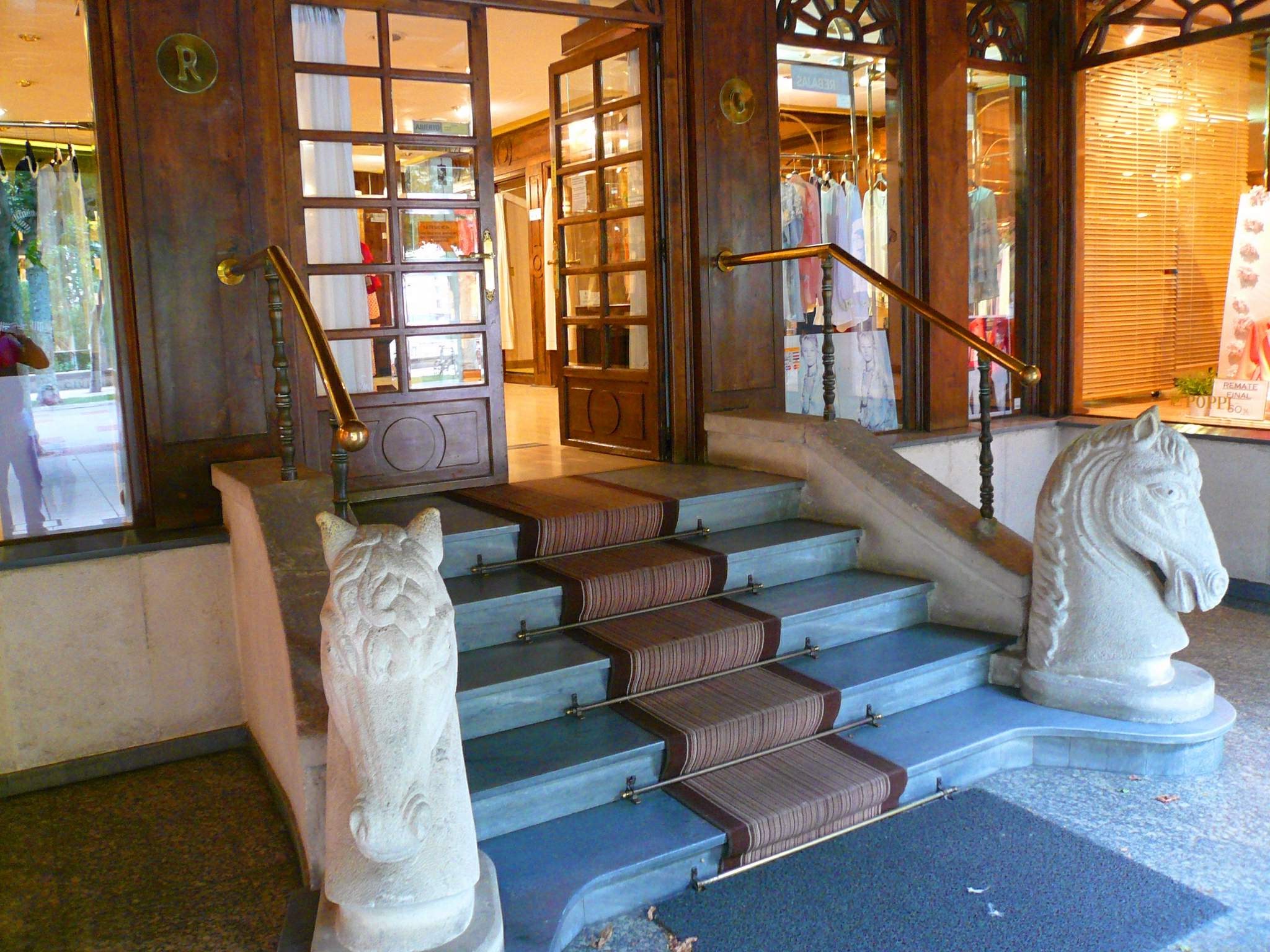 Zwei steinerne Pferdestatuen flankieren den Teppichboden vor einem Geschäft mit Glastüren, Schaufensterpuppen in Kleidern und Innenbeleuchtung.