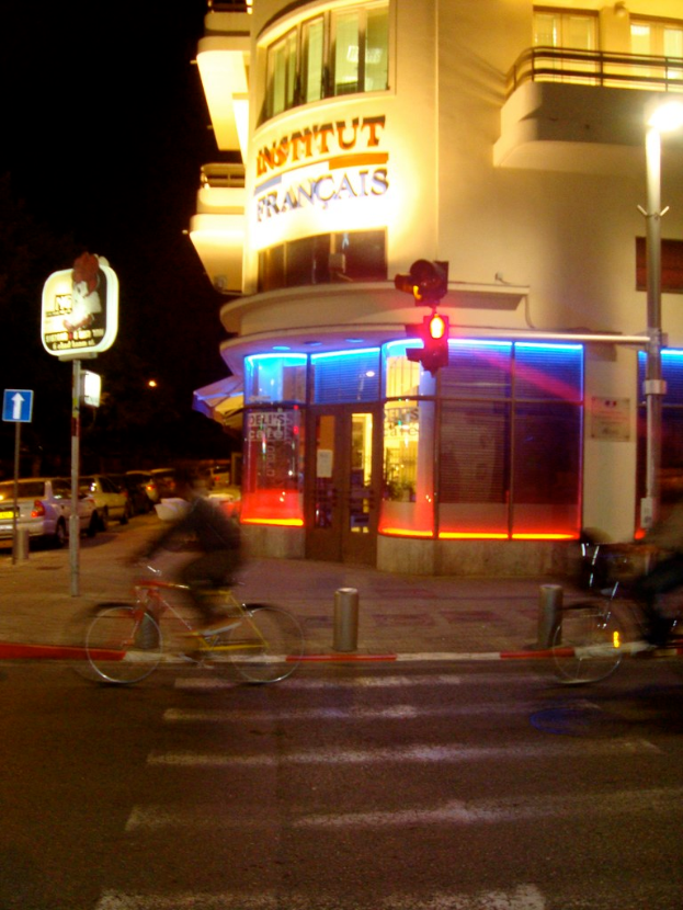 Ein Gebäude mit Text darauf befindet sich auf der rechten Seite des Bildes, mit Pfählen, Lampen, einer Ampel und Schildern mit Text in der Nähe. Es gibt eine Tür am Gebäude. Unten gibt es eine Straße mit zwei Fahrradfahrern. Autos sind auf der linken Seite des Bildes zu sehen.