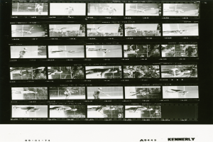 Ein Schwarz-Wei├č-Foto eines Mannes in einem wei├čem Hemd und Shorts, der einen Tennisrahmen in seiner rechten Hand h├Ąlt und einen konzentrierten Ausdruck hat, vor einem Filmstreifen-Hintergrund.