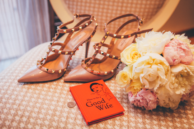 Ein Tisch zeigt ein Buch mit dem Titel "Wie man eine gute Ehefrau ist", einen Blumenstrauß und ein Paar Valentino-Schuhe, vor einem Vorhanghintergrund.