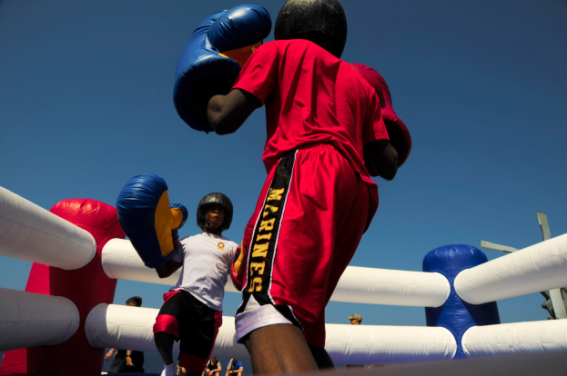 Zwei Boxer in einem Boxring, einer in rotem Hemd und blauen Shorts, kämpfen mit Handschuhen, umgeben von ein paar Menschen und unter einem klaren blauen Himmel.