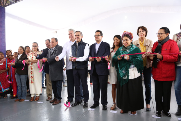Eine Gruppe von Menschen, die lachend eine Schere halten und vor einer Menge stehen, mit einer Säule links daneben, verstreuten Papieren auf dem Boden und einem weißen Tuch im Hintergrund.