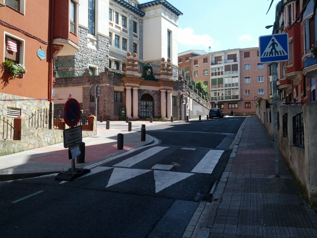 Ein Fahrzeug fährt auf einer Straße mit Verkehrszeichen, Gebäuden mit Fenstern und Pflanzen sowie Säulen.