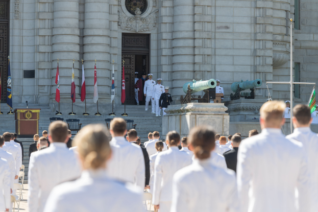 Eine Gruppe von Menschen in Marineuniformen steht vor einem Gebäude mit Säulen und Treppen, mit Flaggen, einem Podium und Kanonen im Hintergrund.